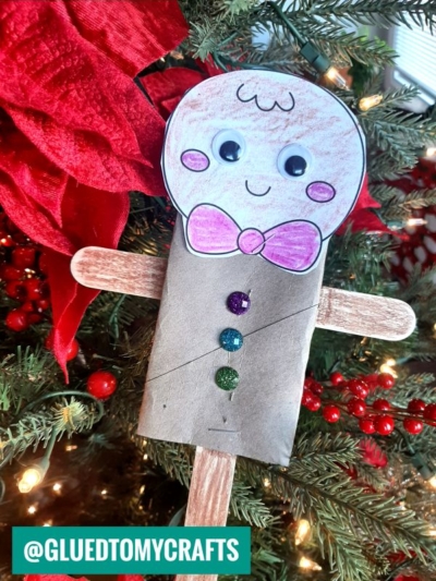 A handmade toilet roll gingerbread man ornament made from a cardboard tube, colored paper, googly eyes, popsicle stick arms, and glitter buttons hangs on a Christmas tree with red ribbon and berries.