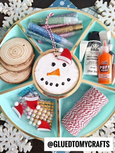 A tray holds supplies for wood slice snowmen—glitter tubes, paints, gems, mini Santa hats, red-and-white twine—with a finished snowman ornament surrounded by snowflake decorations. @GLUEDTOMYCRAFTS is labeled at the bottom.