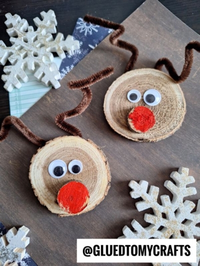 Two handmade wood slice reindeer ornaments with googly eyes, red noses, and brown pipe cleaner antlers are displayed on a surface with decorative white snowflakes nearby. @GLUEDTOMYCRAFTS is written at the bottom.