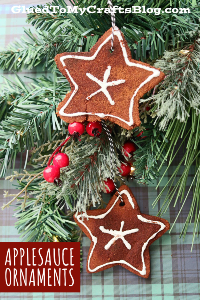 Two star-shaped applesauce ornaments with white icing details hang from a pine branch decorated with red berries. The text at the bottom reads APPLSAUCE ORNAMENTS.