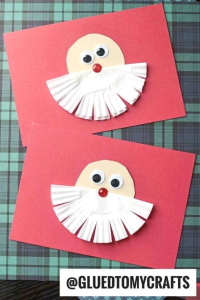 Two handmade Santa cards feature a round face, googly eyes, red nose, and a fun cupcake liner Santa beard. Both are arranged on red paper against a green and blue plaid background. Text @GLUEDTOMYCRAFTS at the bottom.