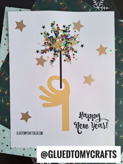 A handmade New Year’s card features a hand holding a paper firework sparkler made of glitter and star confetti, surrounded by gold stars, with the message “Happy New Year!” printed at the bottom.