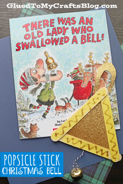 A children's book titled There Was an Old Lady Who Swallowed a Bell! is shown next to a festive popsicle stick Christmas bell craft made of popsicle sticks, gold glitter, and a hanging jingle bell.