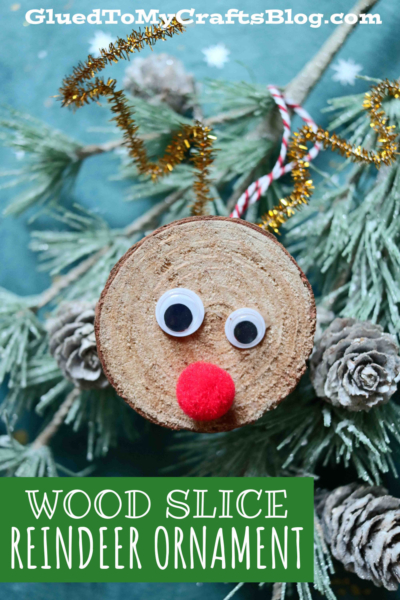 A charming wood slice reindeer ornament with googly eyes, a red pom-pom nose, and gold pipe cleaner antlers hangs from a pine branch adorned with pinecones. Text reads WOOD SLICE REINDEER ORNAMENT.