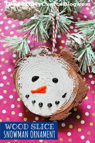 A rustic wood slice ornament painted as a snowman’s face with black eyes, carrot nose, and a dotted smile hangs from a snowy pine branch with pinecones on a red polka-dotted background—perfect for fans of wood slice snowmen.