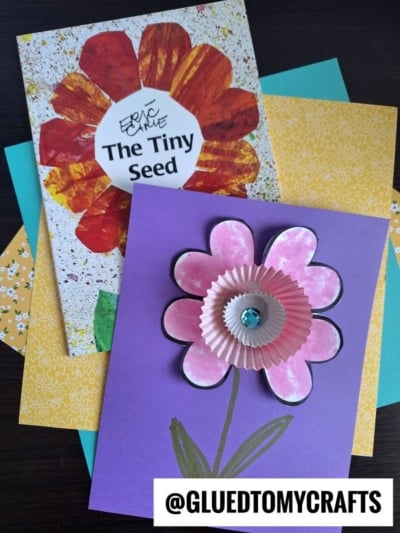 A handmade pink heart flower card with a cupcake liner center lies on colorful paper and the book The Tiny Seed by Eric Carle. The handle @GLUEDTOMYCRAFTS is printed at the bottom.