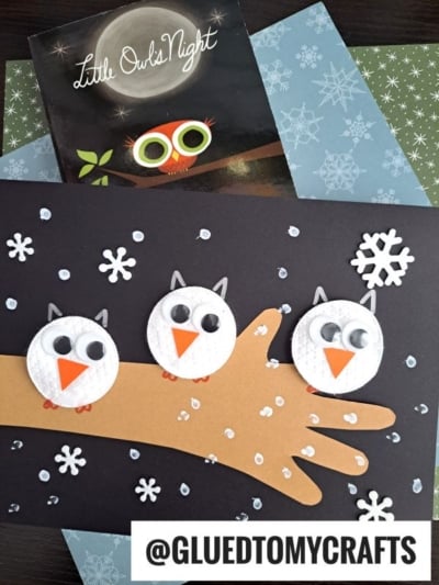 Craft of three white cotton pad owl creations with googly eyes and orange beaks perched on a brown paper branch, decorated with paper snowflakes on a black background. A Little Owls Night book is in the background. @GLUEDTOMYCRAFTS.