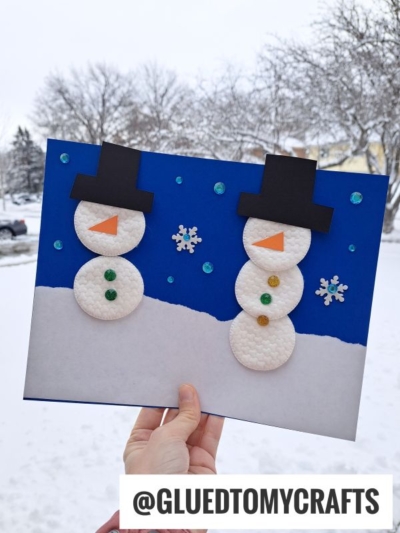 A hand holds a winter craft featuring cotton pad snowmen with paper hats, noses, and buttons on a blue background decorated with snowflakes and sequins, plus snowy trees behindāperfect for making your own cotton snowman puppet.