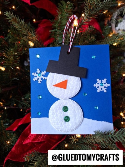 A handmade cotton pad snowman puppet ornament featuring foam circles, a black hat, green buttons, a carrot nose, and snowflake decorations on a blue background hangs on a Christmas tree. Text at the bottom reads @GLUEDTOMYCRAFTS.