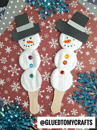 Two cotton pad snowman puppets, each with black paper hats, colored gem buttons, and popsicle stick handles, are displayed on a snowflake-patterned background with blue decorative accents.