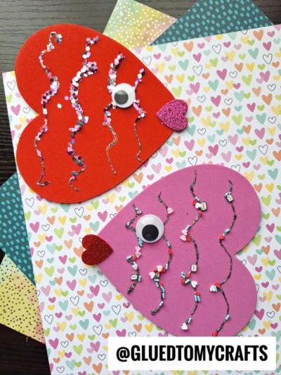 Two fish crafts made from red and pink heart-shaped paper, perfect for a Valentine fish craft for kids, are decorated with wiggly eyes, glitter, small beads, and heart-shaped mouths on colorful papers. @GLUEDTOMYCRAFTS is written at the bottom.