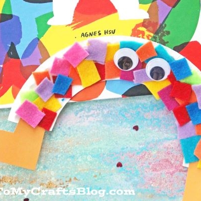A colorful paper plate rainbow with two tan legs, covered in multicolored felt squares and topped with googly eyes, sits on a pastel textured surface next to a bright book cover.