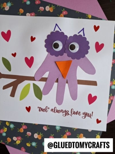A children’s craft features a purple handprint owl with googly eyes and an orange beak, perched on a branch among red and pink hearts. This adorable “owls always love you keepsake” reads: Owl always love you!.