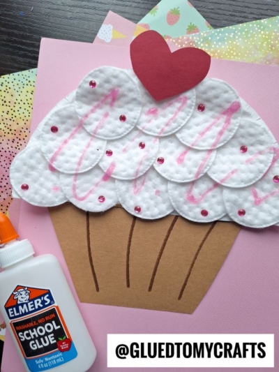 Cupcake craft made from brown and pink paper, cotton pads for icing, pink glitter, a red heart topper, and school glue on a table. This adorable cotton pad cupcake craft features the handle @GLUEDTOMYCRAFTS at the bottom.