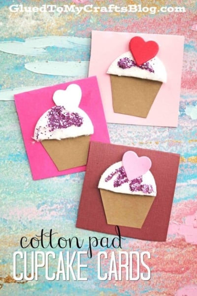 Three handmade cotton pad cupcake cards crafted from cotton pads, paper, and glitter, each topped with a foam heart, are displayed on a colorful painted surface. Text reads cotton pad CUPCAKE CARDS—a delightful cotton pad cupcake craft for all ages.