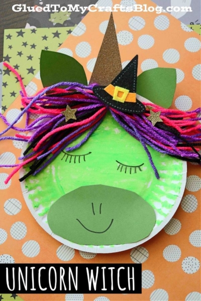 A green paper plate unicorn face with closed eyes, colorful yarn hair, a black witch hat adorned with stars, and green ears, set on decorative orange paper. Text at the bottom reads UNICORN WITCH.