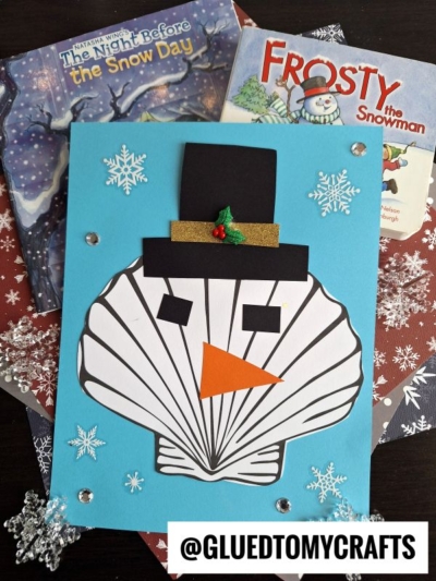 A handmade seashell snowman crafted from white, seashell-shaped paper is glued to a blue card, adorned with snowflakes and gems, and placed atop winter-themed books The Night Before the Snow Day and Frosty the Snowman.