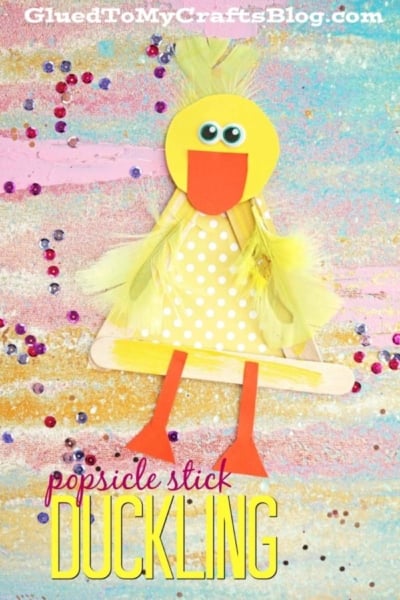 A popsicle stick duckling craft with a yellow polka-dot body, googly eyes, orange beak and feet, and yellow feathers is displayed on a colorful glittery background with scattered sequins—perfect for creating your own adorable popsicle stick duck.