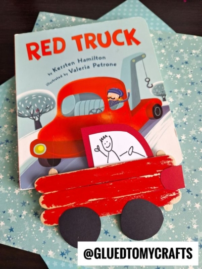 A children’s book titled Red Truck lies on a blue star-patterned surface. In front is a popsicle stick truck craft made of sticks and paper, featuring a stick figure driver. @GLUEDTOMYCRAFTS is written at the bottom.