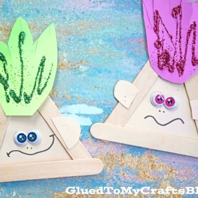 Colorful popsicle stick trolls feature paper faces, googly eyes, bright smiles, and sparkly hair in green and purple. They sit against a blue and pink painted surface for a playful look.
