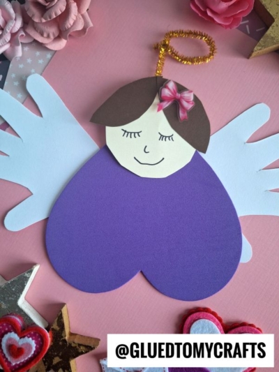 A charming craft foam valentine angel with white wings, a purple heart-shaped body, a sweet smile, brown hair, pink bow, and gold pipe cleaner halo sits on a pink background with decorative items.