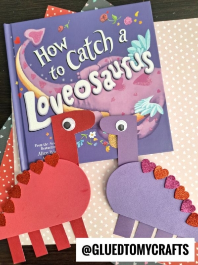 A purple children’s book titled How to Catch a Loveosaurus rests on heart-patterned paper, with two craft foam valentine dinosaurs—one red and one purple—adorned with googly eyes and glittery heart shapes.