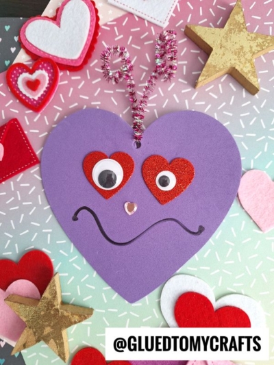 A purple heart-shaped valentine love monster with googly eyes, red heart accents, a wavy smile, pink pom-pom nose, and sparkly pipe cleaner antennae sits among felt hearts and stars on a pastel sprinkle background.