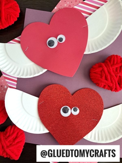 Two heart-shaped crafts with googly eyes and white paper plate wings create a charming Heart With Paper Plate Wings display on colorful paper, surrounded by yarn hearts. @GLUEDTOMYCRAFTS is written at the bottom.