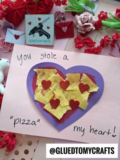 A handmade heart valentine card features a purple heart with yellow crinkled paper and red glitter hearts on pink paper. Text reads “You stole a ‘pizza’ my heart!” Flowers and envelopes decorate the background. @GLUEDTOMYCRAFTS is at the bottom.
