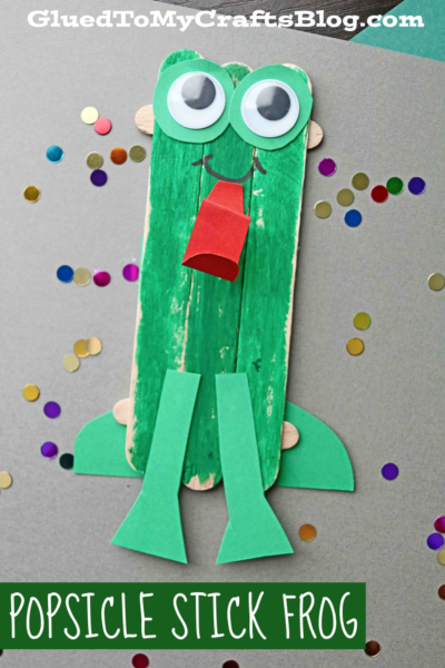 A popsicle stick frog craft made from green-painted sticks with big googly eyes, a red paper tongue, and green feet, surrounded by vibrant confetti. Text reads Popsicle Stick Frog.