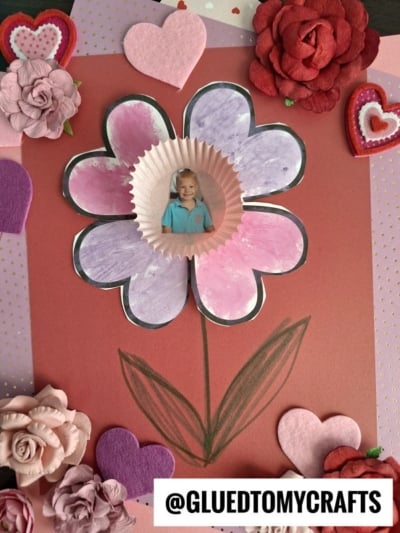A handmade heart flower card featuring a large bloom with pink and purple petals, a child’s photo in the center, and decorated with felt and paper hearts and flowers. Text at the bottom reads @GLUEDTOMYCRAFTS.