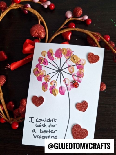A handmade thumbprint valentine card with a dandelion made of pink and gold fingerprints, red glitter hearts, and a tiny ladybug sticker. The card reads, I couldn’t wish for a better valentine. A perfect valentine card kid craft!.