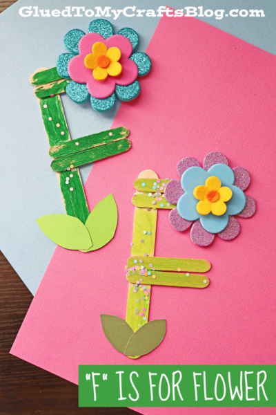 Two craft flowers made from colored popsicle sticks, foam shapes, and paper leaves form the letter F on pink and blue backgrounds. The image caption reads: "F" is for Flower.