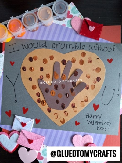 A handmade thumbprint valentine card features a brown paper heart with handprints and sequin “chocolate chips,” reading, “I would crumble without you! Happy Valentine’s Day!” Felt hearts and envelopes decorate the card.