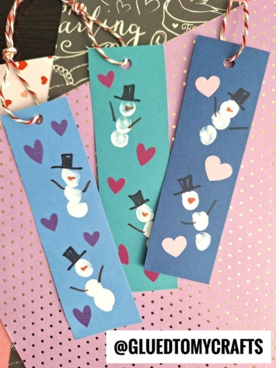 Three handmade thumbprint snowman bookmarks with painted snowmen, colorful hearts, and twine ties are arranged on polka dot and heart-patterned paper. The bookmarks are blue, teal, and purple. @GLUEDTOMYCRAFTS appears at the bottom.