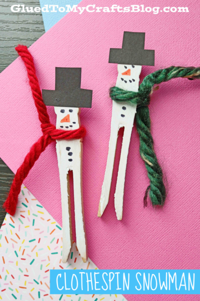 Two wooden clothespins painted white are decorated as Clothespin Snowmen with black paper hats, yarn scarves, and drawn faces. They sit on colorful paper backgrounds featuring the text CLOTHESPIN SNOWMAN.