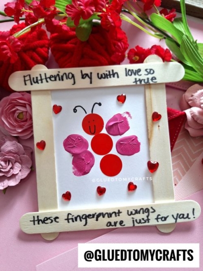 A handmade craft features a ladybug with red paper circles and pink fingerprint wings, framed with popsicle sticks. This charming fingerprint valentine card says, Fluttering by with love so true, these fingerprint wings are just for you!.