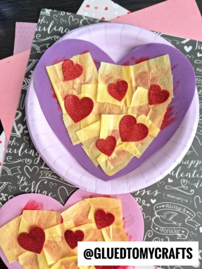 Two heart-shaped crafts made with yellow tissue paper squares and red glitter hearts, resembling a heart valentine pizza, are displayed on purple and pink plates. The background includes Valentine-themed paper and the text @GLUETOMYCRAFTS.