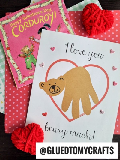 A handmade Valentines card features a bear made from a brown cut-out handprint inside a pink heart, with the text Love You Beary Much! Surrounded by small heart decorations, yarn hearts, and a Corduroy book.