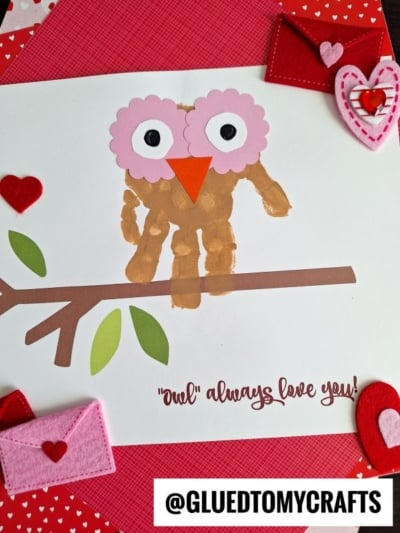A handmade card features an owl made from a painted handprint with pink paper eyes and an orange beak, sitting on a branch. Decorated with felt hearts and envelopes, this is the perfect owls always love you keepsake!.
