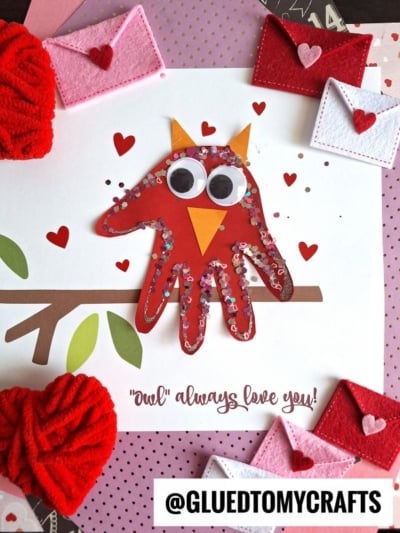 A handmade owl card made from a red handprint with googly eyes, an orange beak, and heart-shaped decorations. Surrounded by felt heart envelopes and red yarn, it makes a sweet "owls always love you keepsake.