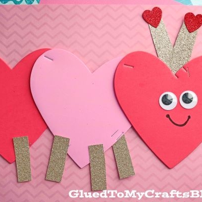 Three heart-shaped paper cutouts form a charming heart shaped caterpillar, featuring red heart antennae, googly eyes, a smile, and glittery gold legs, arranged on pink and blue patterned paper.