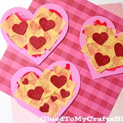 A heart valentine pizza craft made with pink paper, yellow squares as cheese, red sauce, and glittery red heart toppings, displayed on a pink plaid background.