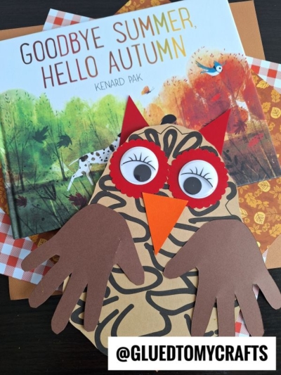 A children’s book titled Goodbye Summer, Hello Autumn sits on a table with a handmade paper pinecone owl craft made from brown, orange, and red paper, featuring handprint wings and large googly eyes.