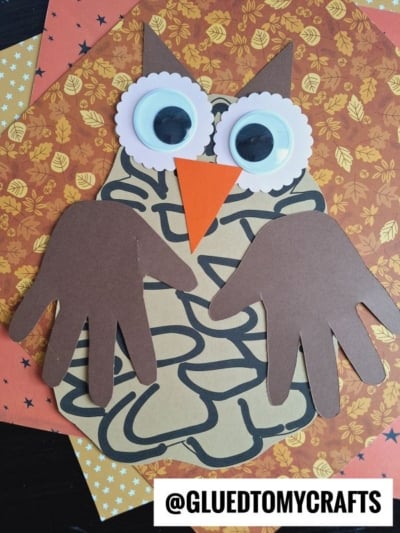 A handmade paper pinecone owl craft made from brown paper cutouts, featuring handprint wings, large googly eyes, and an orange beak, displayed on autumn-patterned paper. Text reads @GLUEDTOMYCRAFTS.