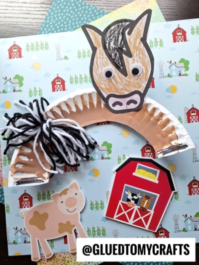 A paper plate horse craft made with googly eyes and black-and-white yarn for the mane sits on farm-themed paper featuring barns and cows. The handle @GLUEDTOMYCRAFTS is visible in the scene.