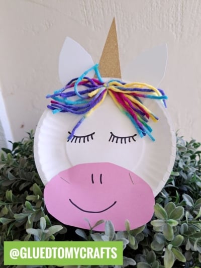 A whimsical paper plate unicorn craft features a gold horn, colorful yarn mane, closed eyes, and a pink paper snout, resting on green foliage. A green label reads @GLUEDTOMYCRAFTS—perfect inspiration for your next paper unicorn project!.