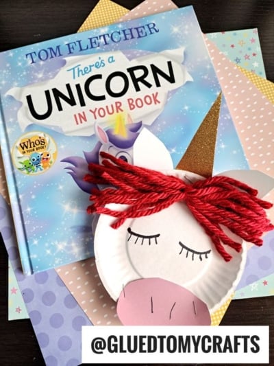 A children’s book titled Theres a Unicorn in Your Book by Tom Fletcher is shown with pastel-colored papers and a paper plate unicorn craft with red yarn hair. A whimsical paper unicorn comes to life, with @GLUEDTOMYCRAFTS at the bottom.