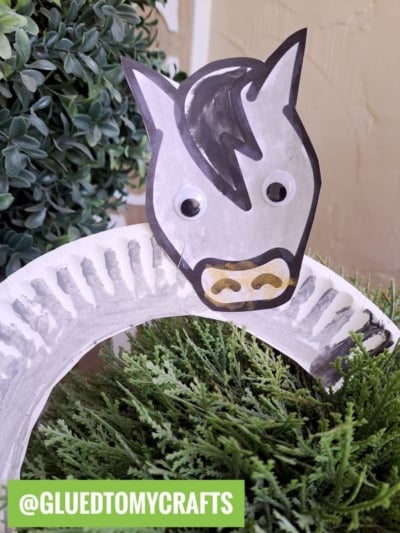 A paper plate craft shaped like a zebra or paper plate horse, featuring a zebra face with googly eyes and a black mane, set among green artificial plants. The handle @GLUEDTOMYCRAFTS is displayed in the bottom left corner.