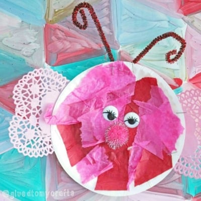A paper plate lovebug craft butterfly with pink and red tissue paper, googly eyes, a red pompom nose, pink doily wings, and red pipe cleaner antennae, set against a colorful painted background.